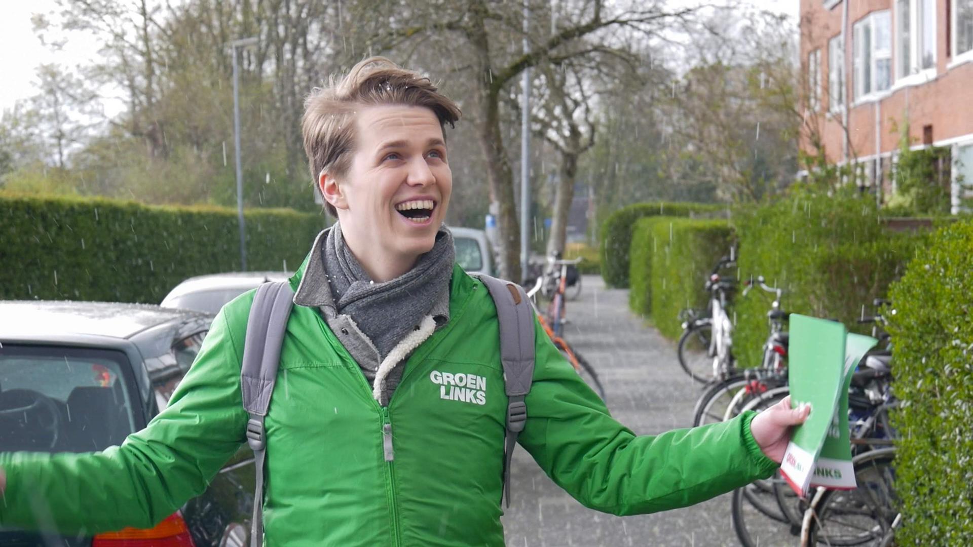 Een GroenLinks-vrijwilliger kijkt blij terwijl hij in de regen flyers vasthoudt